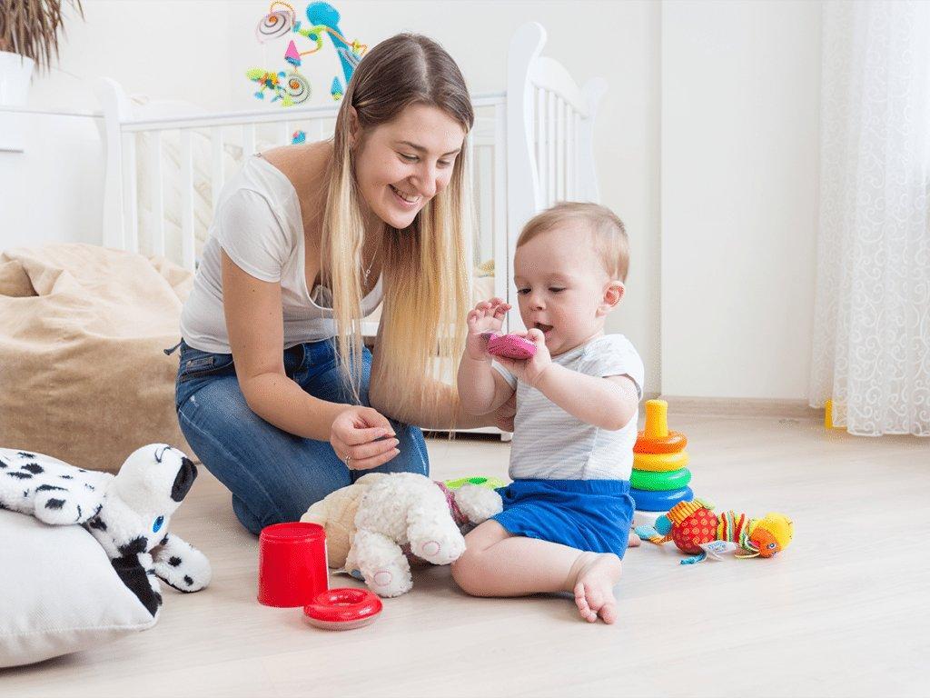 A caregiver sits on the floor with a toddler surrounded by stuffed animals, stacking rings, and plastic cups. The setting is the kind of natural play context NDBI models are built around: the therapist or trained parent follows the child’s interest, uses the toy the child has already chosen, and reinforces communication and play with materials the child finds intrinsically motivating.