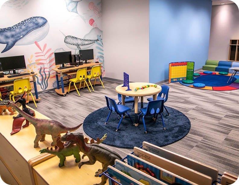 An Akoya Behavioral Health clinic features an ocean-themed mural, child-sized workstations, a group activity table, and a sensory-friendly reading area with toys and books.